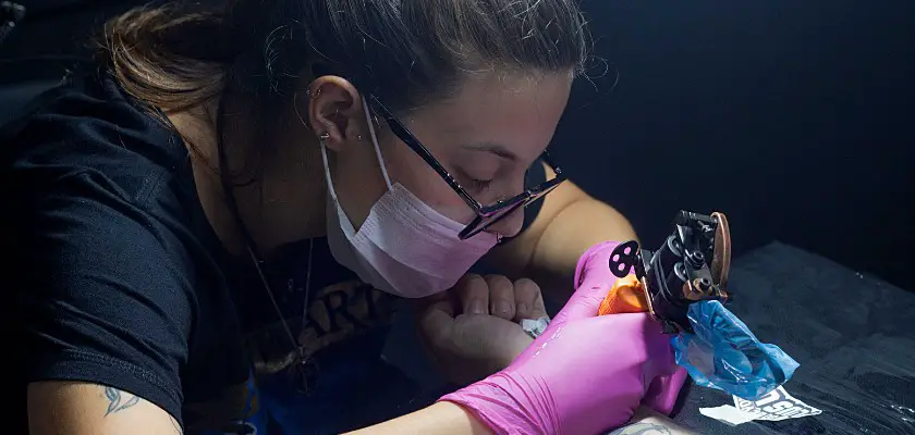 Tatuatore donna con la mascherina che effettua un tatuaggio sul braccio del suo cliente