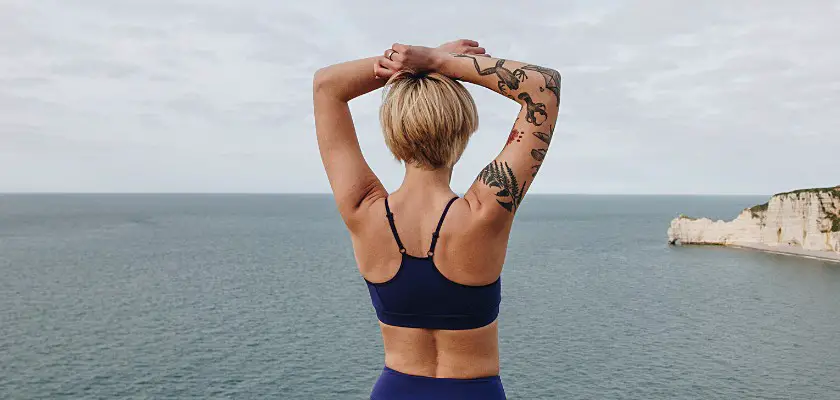 Vista posteriore di una ragazza bionda atletica con le braccia sopra la testa mentre osserva il mare – Dopo quanto posso esporre il tatuaggio al sole