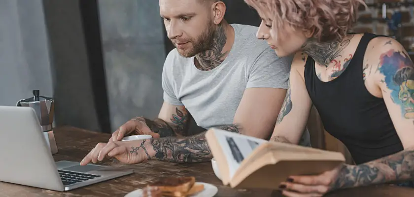 Coppia tatuata seduti a tavola in cucina mentre utilizzano il laptop – Psicologia dei tatuaggi