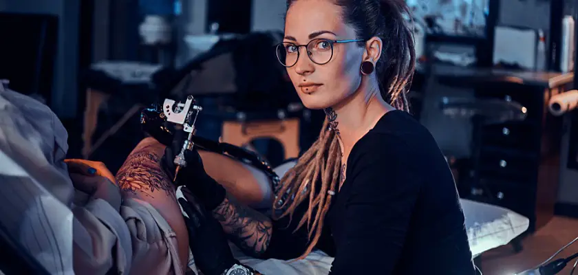 Bellissima tatuatrice con gli occhiali mentre sta effettuando un nuovo tatuaggio sulla gamba destra della sua cliente