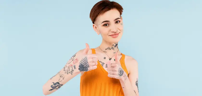 Bella ragazza tatuata con i capelli corti che guarda la fotocamera mentre fa il gesto dell’Ok con entrambi i pollici delle mani